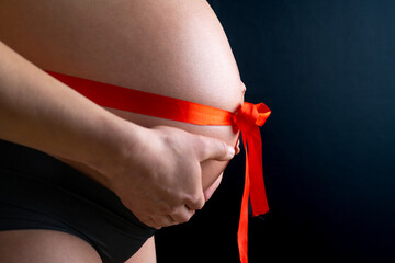 Side closeup of naked round belly with a tied red bow. Last month of pregnancy - week 36. Cropped. Black background.