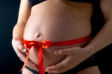 Closeup of naked round belly with a tied red bow. Last month of pregnancy - week 36. Cropped, front view. Black background.