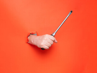 Hand holding a big screwdriver through torn orange paper. No face, concept.
