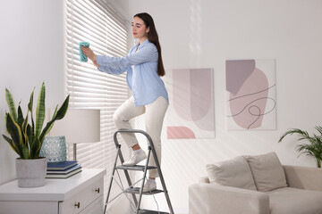 Woman on metal ladder wiping blinds at home