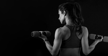 Fototapeta na wymiar Female sporty muscular young serious woman doing strength workout on the shoulders, biceps and arms in sport bra holding dumbbells on black background. Closeup