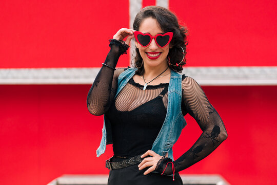 retrato de bella mujer latina sonriendo con maquillaje jugando con sus lentes rojos al aire libre en un fondo rojo estilo 80s 90s