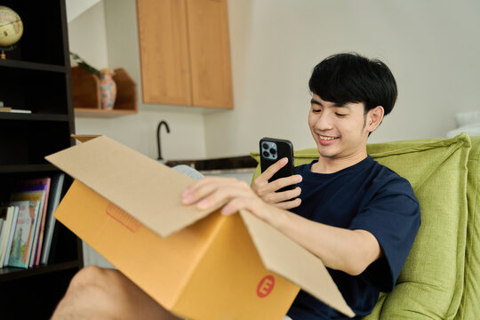 Young asian man unpacking  awaited parce looking inside, sitting at sofa, satisfied happy customer opening cardboard box with online store order, good shipping delivery service