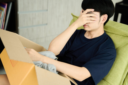 Young asian man unpacking awaited parce looking inside, sitting at sofa, unsatisfied customer opening cardboard box with online store order ,Delivery service concept.