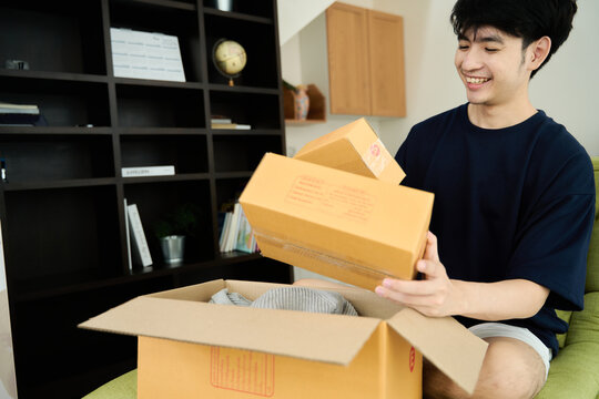 Young asian man unpacking  awaited parce looking inside, sitting at sofa, satisfied happy customer opening cardboard box with online store order, good shipping delivery service
