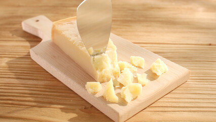 Hard cheese is chipped off with a knife on a cutting board. Wooden background. Parmesan with broken pieces. Traditional Italian cheese grana parmigiano reggiano. Close-up