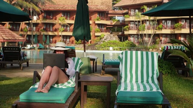 Woman freelancer works online browsing in internet on laptop sits in sunbed on tropical resort near hotel on vacation. Brunette entrepreneur middle aged female. Remote work distant job freelance.