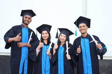 Smiling students in academic caps and gowns celebrating graduation holding certificates and showing thumbs up