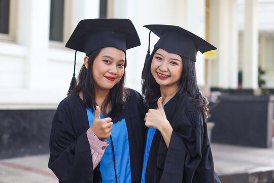 Two Asian women giving thumbs up in university celebrating graduation day with happiness after receiving university degree certificate