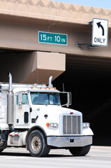 A white, heavy-duty commercial truck transporting materials at a freeway underpass, where vehicle maximum height and 'turn only' traffic signs are visible