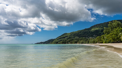 A wave of turquoise ocean rolls to the shore and spreads on the beach. Wet sand glistens. A hill overgrown with tropical vegetation, against a background of blue sky and clouds. Seychelles. Mahe.
