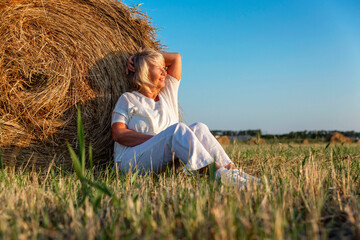 Smiling elderly woman in a white sweater and trousers in a field with hay rolls at sunset. Farming,...