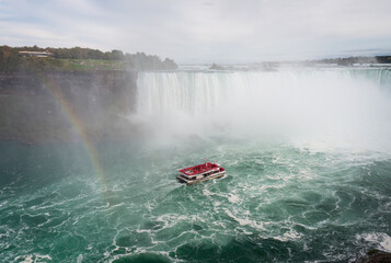 Fototapeta premium Rainbows and cruising boat at Niagara Falls. Tourists wearing pink raincoats on the boat. Canada.