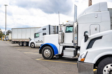 Different make and models of white big rig semi trucks with semi trailers standing in row on truck stop parking lot for truck driver rest