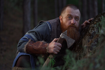 Lurking medieval red-haired viking warrior with beard with an ax in forest