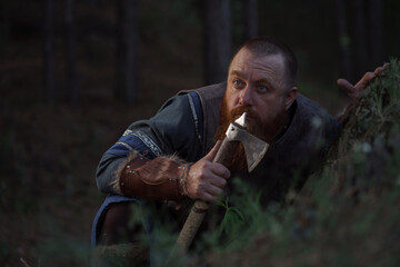 Lurking medieval red-haired viking warrior with beard with an ax in forest