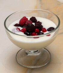 a glass bowl with liquid yogurt and forest fruits: blueberries, blackberries, blackthorns, on a marble table
