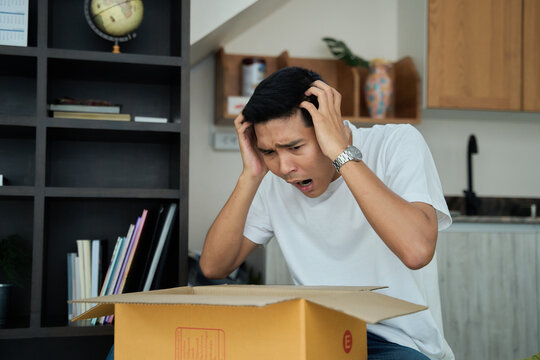 Young asian man unpacking awaited parce looking inside, sitting at sofa, unsatisfied customer opening cardboard box with online store order ,Delivery service concept.