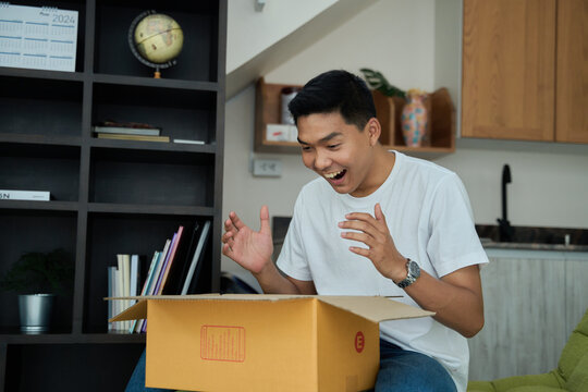 Young asian man unpacking  awaited parce looking inside, sitting at sofa, satisfied happy customer opening cardboard box with online store order, good shipping delivery service