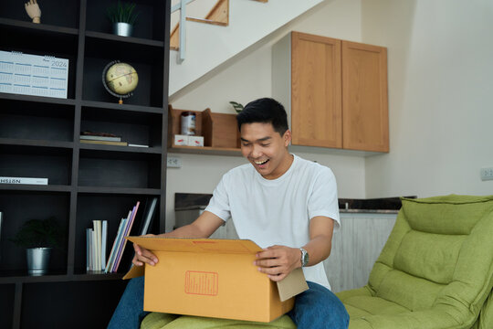 Young asian man unpacking  awaited parce looking inside, sitting at sofa, satisfied happy customer opening cardboard box with online store order, good shipping delivery service