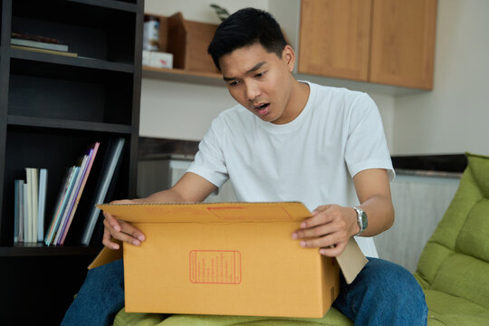 Young asian man unpacking awaited parce looking inside, sitting at sofa, unsatisfied customer opening cardboard box with online store order ,Delivery service concept.