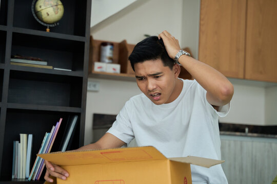 Young asian man unpacking awaited parce looking inside, sitting at sofa, unsatisfied customer opening cardboard box with online store order ,Delivery service concept.