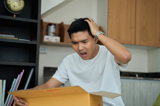 Young asian man unpacking awaited parce looking inside, sitting at sofa, unsatisfied customer opening cardboard box with online store order ,Delivery service concept.