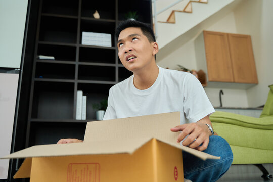 Young asian man unpacking awaited parce looking inside, sitting at sofa, unsatisfied customer opening cardboard box with online store order ,Delivery service concept.