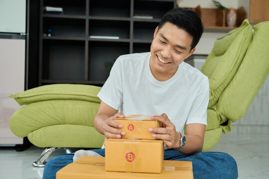 Young asian man unpacking  awaited parce looking inside, sitting at sofa, satisfied happy customer opening cardboard box with online store order, good shipping delivery service