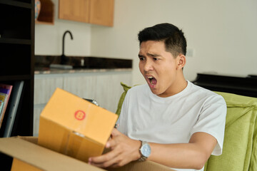 Young asian man unpacking awaited parce looking inside, sitting at sofa, unsatisfied customer opening cardboard box with online store order ,Delivery service concept.