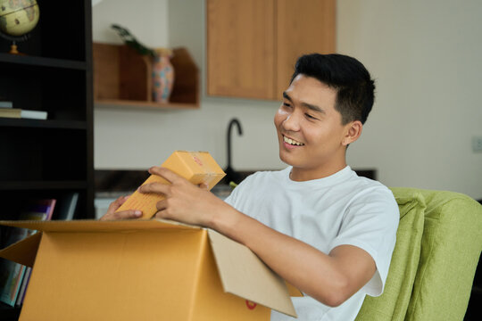 Young asian man unpacking  awaited parce looking inside, sitting at sofa, satisfied happy customer opening cardboard box with online store order, good shipping delivery service