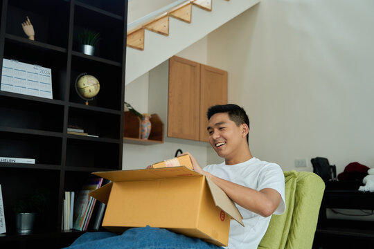 Young asian man unpacking awaited parce looking inside, sitting at sofa, unsatisfied customer opening cardboard box with online store order ,Delivery service concept.