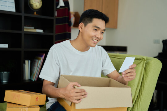 Young asian man unpacking  awaited parce looking inside, sitting at sofa, satisfied happy customer opening cardboard box with online store order, good shipping delivery service