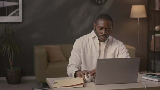 Waist up of young Black businessman playing with baseball while working on laptop computer at desk in home office