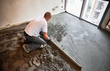 Male construction worker using screed rail while screeding floor in living room. Man flattening and...