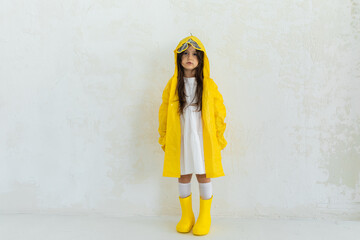 Sad disappointed cute little girl is wearing yellow waterproof raincoat, rubber boots and white dress. Weather forecast concept. space for text