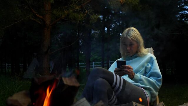 A young woman using a mobile phone, sitting by a burning campfire in a camp at dusk. A female travel blogger enjoys the warmth and comfort by the fire, communicates with subscribers on her smartphone.