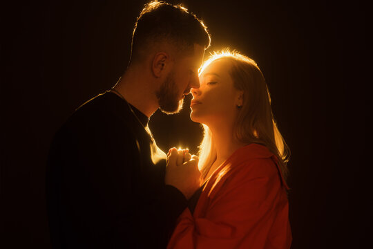 Side view of young bearded man and blond haired woman holding hands and kissing gently in darkness against glowing warm light. Horizontal photo