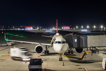An airplane stands on the boarding bridge at night with a moving plane in the background