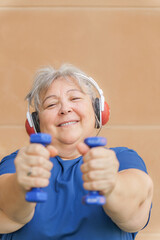 Naklejka premium obese white-haired older woman exercising with dumbbells and headphones listening to music