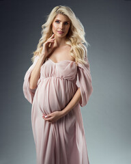 Beautiful Pregnant Woman Portrait in Pink Chiffon Dress over Gray Background. Happy Mother with Curly Long Hair in Silk Gown. Pregnancy Fashion