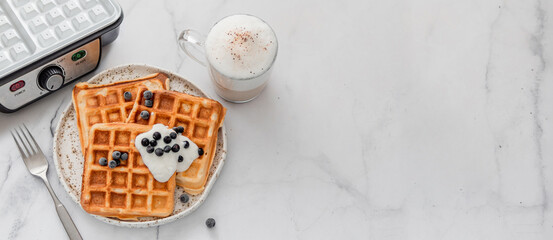 Delicious homemade baked belgian waffles with greek yogurt, blueberries and cappuccino and electric waffle maker with white ceramic coated on white marble background. Copy space