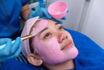 An esthetician applies clay facial mask to a customer's face prior to using a phototherapy device...