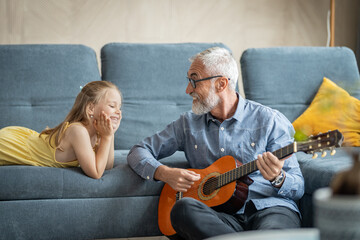 Little girl lying on the sofa and smiling as her grandfather plays guitar
