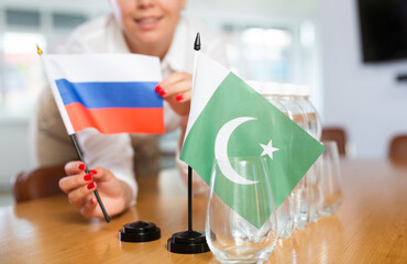 Positive young woman putting little flag of Russia on table next to the flag of Pakistan and...