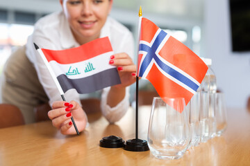 Little flag of Norway on table with bottles of water and flag of Iraq put next to it by positive young woman in meeting room