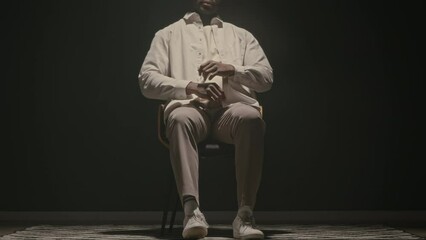 Cropped stab studio portrait of Black man in white clothes sitting down on wooden chair in middle of room on clear black background with dimmed spotlight shining from above