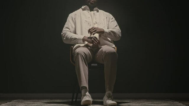 Cropped stab studio portrait of Black man in white clothes sitting down on wooden chair in middle of room on clear black background with dimmed spotlight shining from above
