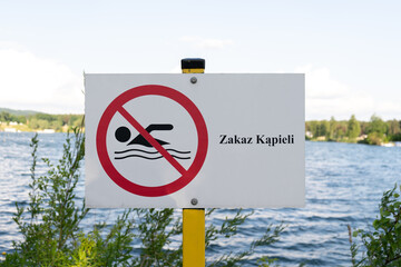 No Swimming sign post plaque, with  water in background. No swim area signage pillar, mounted on the shore of lake in Poland. Text in Polish Zakaz kąpieli, means Swimming prohibited. - Powered by Adobe