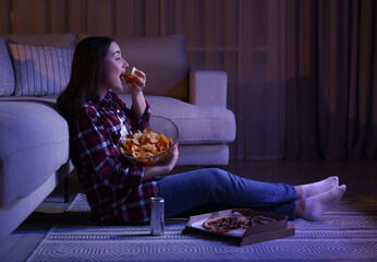 Young woman eating chips while watching TV in room at night. Bad habit